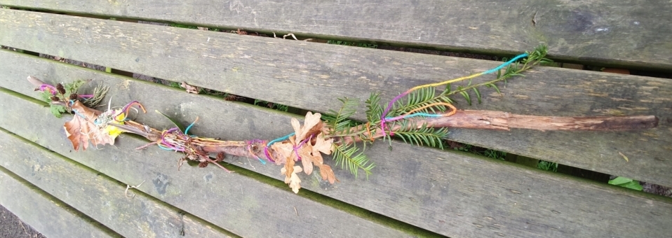 Freddie is in his garden holding up his finished journey stick. There is moss, leaves and towards the bottom, there are bright yellow flowers.