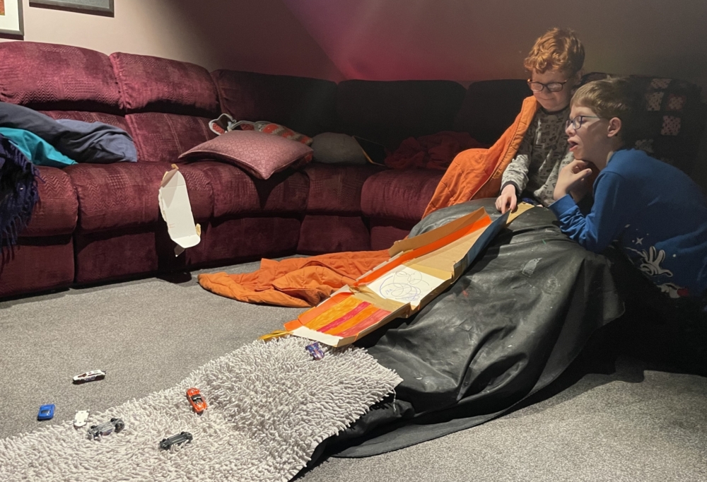 Ryan and his brother are racing cars down their cardboard ramp propped up by cushions. In this photo they have a grey, bobbly matt at the bottom.