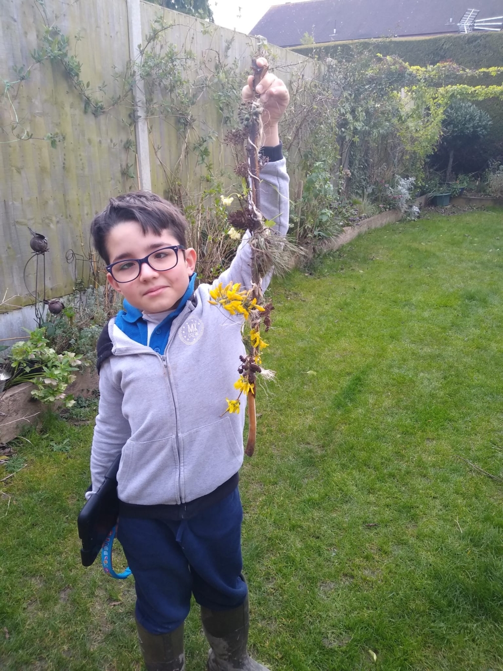 Freddie is in his garden holding up his finished journey stick. There is moss, leaves and towards the bottom, there are bright yellow flowers.