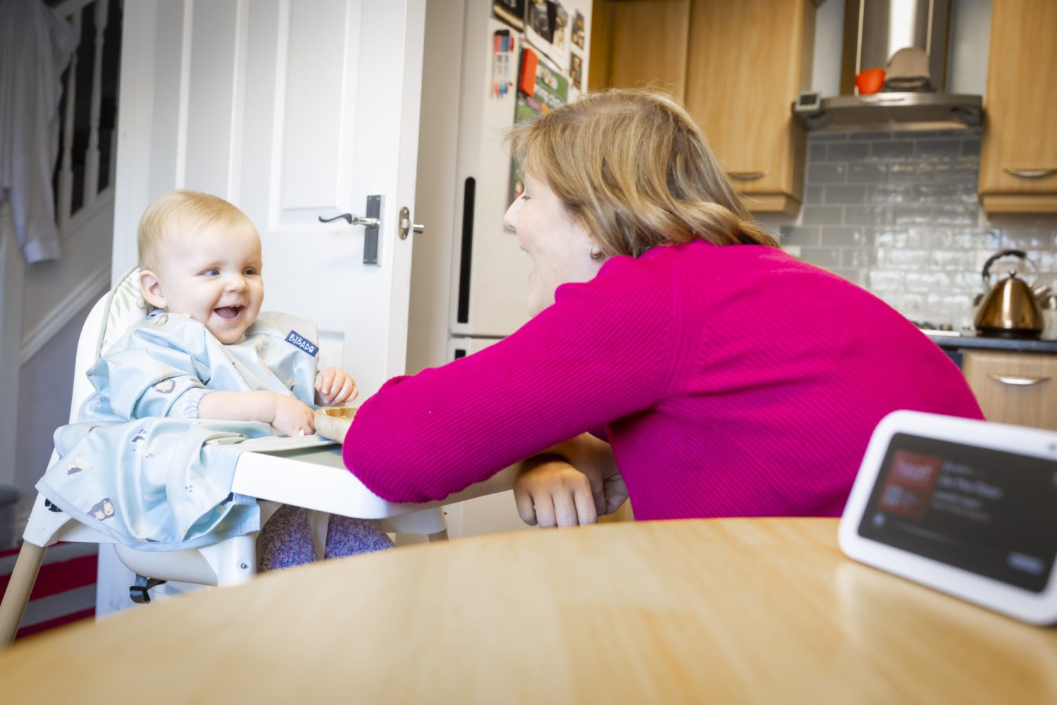 A parent sits with her child in a high chair. They are laughing as they listen to an Echo Show.